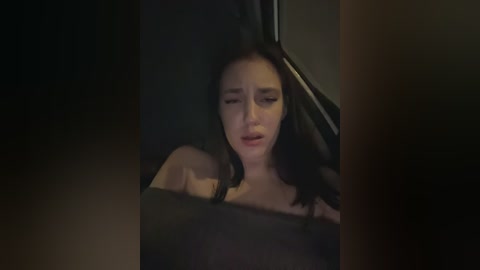 Video of a woman with dark hair and light skin, wearing a black top, sitting in a dimly lit car, looking distressed and crying.