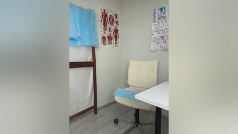 Video of a small, minimalistic medical exam room with a white exam chair, a white desk, and anatomical posters on the walls. A blue exam gown hangs on a wooden stand.
