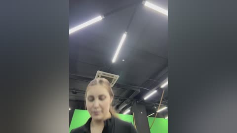 A blurry video of a young woman with light skin and blonde hair, wearing a black blazer, captured in a dimly lit room with green screen in the background and fluorescent lighting.