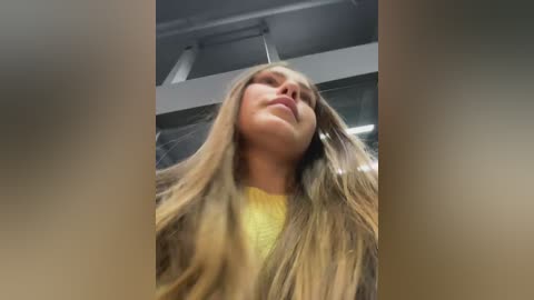 Video of a young woman with long, wavy, light brown hair, wearing a yellow shirt, gazing upward. The background features a modern, industrial ceiling with white beams and fluorescent lights.