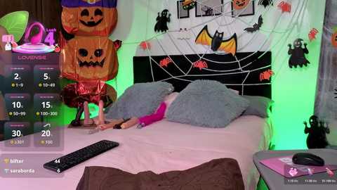 Video of a girl in pink pajamas lying on a bed with a TV remote. Halloween decorations in the background, including a pumpkin, bats, and spider web.
