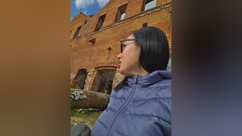 Video of a woman with straight black hair and black-framed glasses, wearing a blue puffer jacket, standing in front of an old, weathered brick building with arched windows and graffiti on a wall.