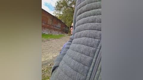 A video featuring a close-up of a person wearing a grey quilted jacket, standing near a brick wall and a grassy area in a park. The background includes trees and a clear sky.