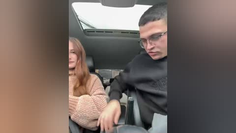 Video of a young Caucasian couple in a car; woman with long brown hair, beige sweater, smiling; man with short dark hair, glasses, black hoodie, frowning.