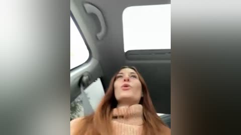 Video of a woman with long brown hair, wearing a beige turtleneck sweater, leaning back in a car, mouth slightly open, surrounded by gray interior and a window.
