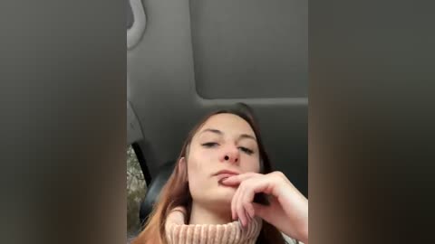 Video of a young woman with light skin, brown hair, and fair complexion, wearing a brown sweater, resting her chin on her hand, seated in a car with gray seats and a patterned floor.