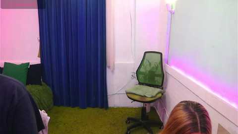Video of a small, dimly-lit room with green carpet, white walls, blue curtains, and a green mesh chair; a person with shoulder-length brown hair is seen from the back.