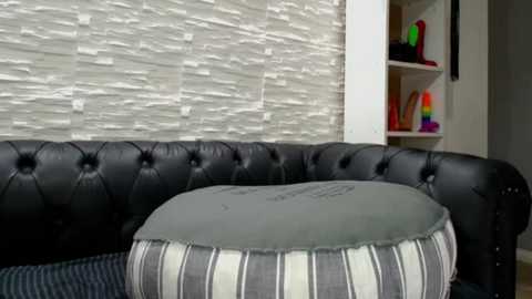 Video of a modern living room with a black tufted leather couch, white textured wall, and a striped gray and white cushioned ottoman. Background includes a colorful toy shelf.