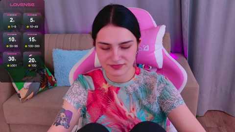 Video of a young woman with light skin and long black hair, wearing a tie-dye shirt, sitting on a pink gaming chair in a cozy living room.