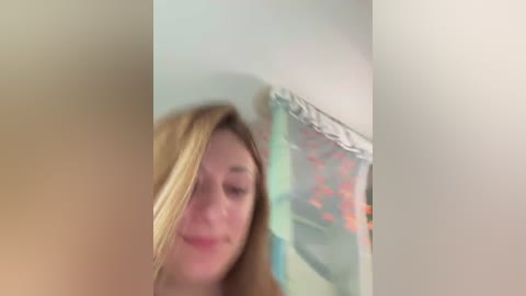 Video of a blonde woman with light skin and a slight smile, partially obscured by a blurry, colorful shower curtain. The background is off-white with soft lighting.