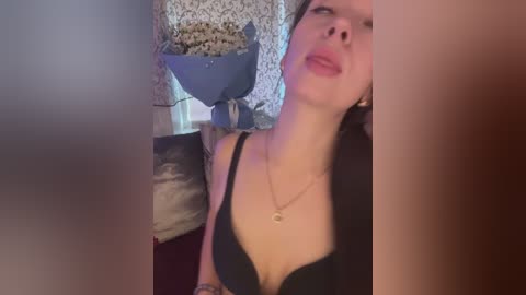 A video of a young woman with fair skin, black hair, wearing a black bra, reclining on a bed with a floral-patterned wallpaper background.