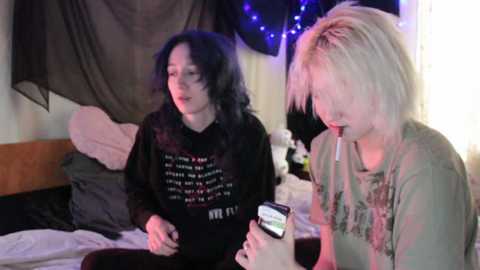 Video of two women with long hair, one in black hoodie, the other in grey shirt, sitting on a bed, holding a cellphone.