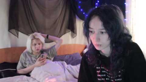 Video of a young woman with long, wavy black hair, wearing a black hoodie, holding a phone. A pale, curly-haired woman in a grey hoodie is in bed, looking at the phone. Background includes a draped curtain and blue fairy lights.