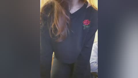 Video of a young woman with long, wavy brown hair, wearing a black sweatshirt with a red rose patch and black leggings, standing indoors against a blurred background.