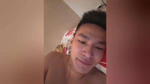 A young Asian man with short black hair, light skin, and a slight smile lies on a bed with a red and white patterned pillow, partially covered by beige walls.