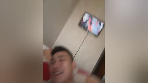 A blurred video of a young man with short dark hair, wearing a red shirt, smiling, in a dimly lit room with a TV displaying a colorful screen above him.