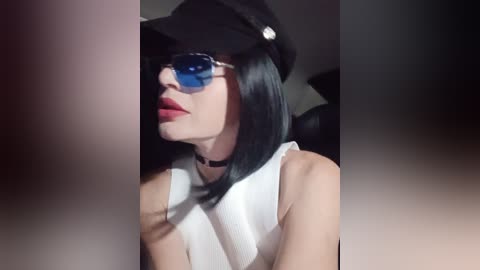 Video of a woman with pale skin, black bob hair, and blue-tinted sunglasses, wearing a white sleeveless top and black hat.