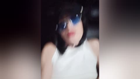 A blurred video shows a young woman with pale skin, wearing a white sleeveless top and dark sunglasses. Her dark hair is partially visible, and her face is obscured by the lens.