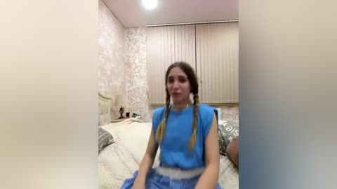 A video of a young girl with long braided hair, wearing a blue top and skirt, sitting on a bed in a patterned wallpaper room.