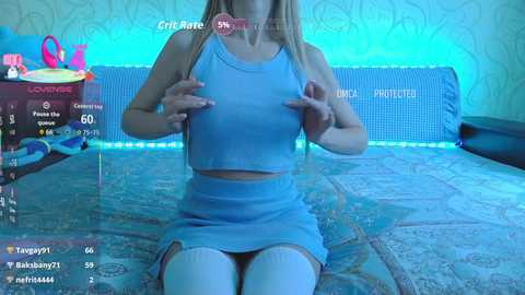 Video of a young, blonde woman with fair skin, wearing a light blue crop top and skirt, sitting on a bed with patterned bedding. Virtual chat interface overlaid on the image.