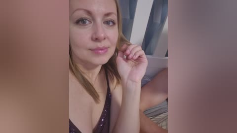 Video of a fair-skinned, blonde woman with medium-length hair, wearing a dark purple sequined bra, lying on a bed with blue curtains in the background.