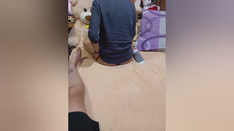 Video of a person sitting on a beige blanket in a bedroom. The person, wearing a navy blue shirt, is backlit. Background includes stuffed animals and a bed with a purple and white striped pillow.