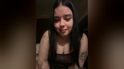 A video of a young, pale-skinned woman with long black hair, wearing a black lace top, sitting indoors. She has a serene expression and visible tattoos on her arms.