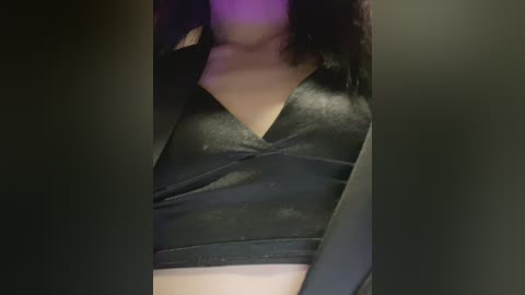 Video of a person with medium skin tone, wearing a black, shiny, V-neck top. The background is blurred, featuring dark, possibly greenish tones. The individual's shoulders and chest are visible, with a hint of purple lighting.