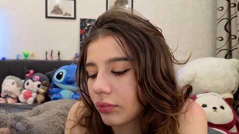 Video of a young girl with long brown hair, wearing a pink shirt, sitting in a cozy room filled with stuffed toys, including Stitch and a teddy bear.