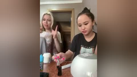 Video of two young women in a bathroom; one has blonde hair, the other dark hair. They're jokingly pointing at each other while holding a white mug and a black bottle.