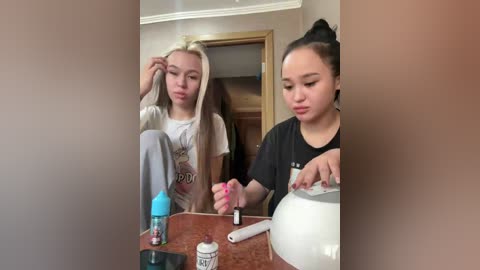 A video of two young women, one with blonde hair in a light t-shirt, the other with dark hair in a black shirt, applying makeup in a bathroom with a wooden door in the background.
