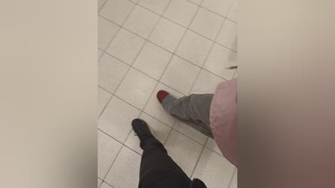 Video of a person's legs, wearing grey pants and black socks with red heels, standing on a tiled floor with white and grey tiles, viewed from above.