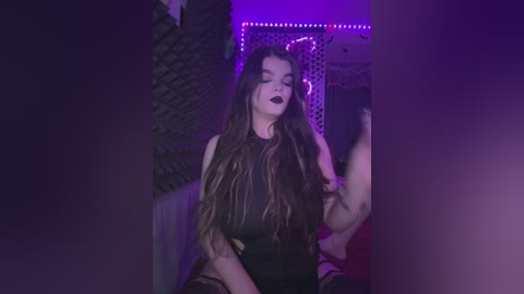 Video of a young woman with long, dark hair, wearing a black, sleeveless dress, seated in a dimly lit, purple-lit room. She has dark makeup and appears contemplative.