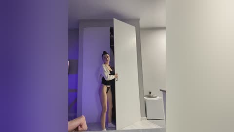 Video of a young, slender woman in a white blouse and black skirt, standing in a dimly lit, minimalist bathroom with a white toilet, a closed door, and a purple accent light.