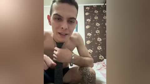 A young, shirtless man with short, spiky hair, wearing a tie, sits on a bed in a retro-themed room with floral wallpaper. He has a tattoo on his left forearm.