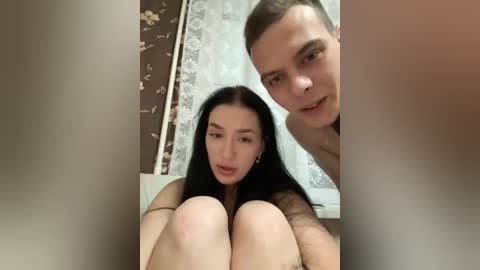 Video of a young woman with long black hair and a fair complexion, sitting on a bed, topless, with a man behind her, both smiling, in a dimly lit bedroom with floral wallpaper.