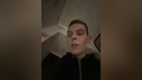 A blurry video shows a young man with short brown hair, wearing a black jacket, standing in a dimly lit room with white walls and a staircase in the background.