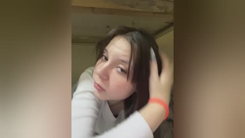 Video of a young woman with fair skin and straight brown hair, wearing a white shirt, sitting in a wooden-framed room, looking contemplative with one hand in her hair.