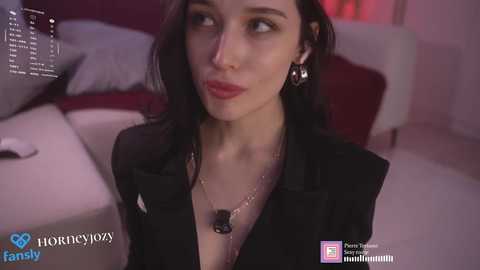 Video of a light-skinned woman with dark hair, wearing a black blazer and a necklace, in a dimly lit room.