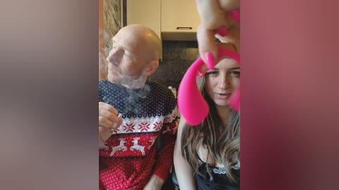 Video of a bald, bearded man wearing a Christmas sweater, smoking a cigarette, and a young woman with long brown hair in a pink bunny costume, indoors with kitchen cabinets in the background.