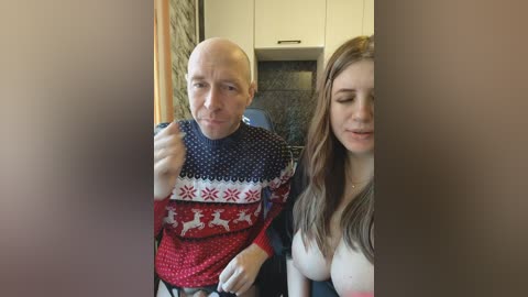 Video of a bald, middle-aged man in a festive sweater, sitting next to a young woman with long brown hair and cleavage, in a modern kitchen.