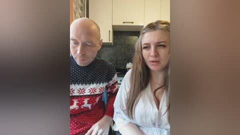 Video of a bald man in a festive sweater and a young woman with long brown hair in a white shirt, seated in a modern kitchen.
