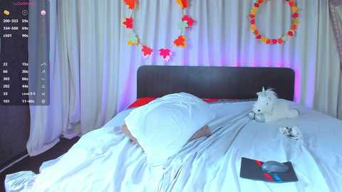 Video of a bed with a person sleeping under white sheets, surrounded by festive decorations including a unicorn plush, a rainbow wreath, and colorful balloons.