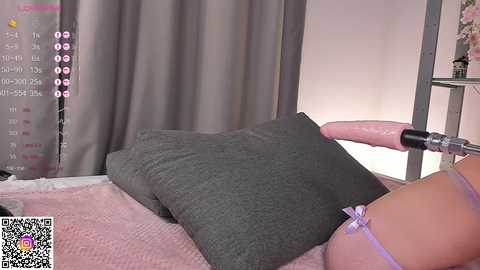 A video of a woman lying on a bed, wearing a grey shirt, with a pink dildo inserted into her vagina, set against a grey curtain background.