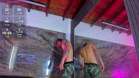 Video of a shirtless man with a tattoo, wearing camo shorts, reflected in a mirror, standing in a dimly lit room with marble walls and red accents.