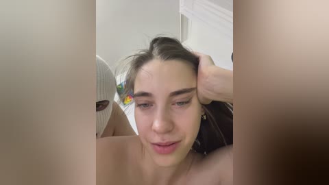 Video of a young woman with fair skin and straight brown hair, wearing a colorful t-shirt, sitting in a brightly lit room, smiling and looking down.