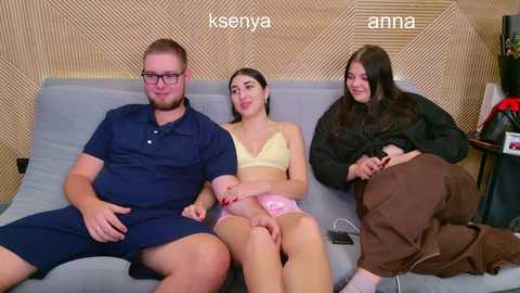 Video of three people on a couch: a bearded man in a blue polo, a woman in a yellow bra, and a woman in a black sweater. Background shows wooden paneling and a bookshelf.