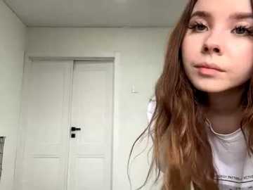 Video of a young woman with fair skin, long wavy brown hair, and a neutral expression, wearing a white t-shirt. Background shows a white door and white wall.