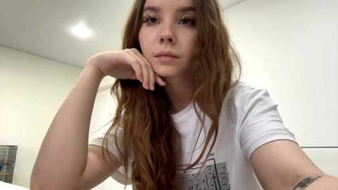 Video of a young woman with long, wavy brown hair, wearing a white t-shirt, resting her chin on her hand, looking contemplative. Background features a white wall and a bed with a plaid blanket.