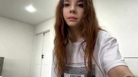 Video of a young woman with long, wavy auburn hair, wearing a white T-shirt with a graphic design, standing in a minimalist, white-walled room with a door and recessed lighting.